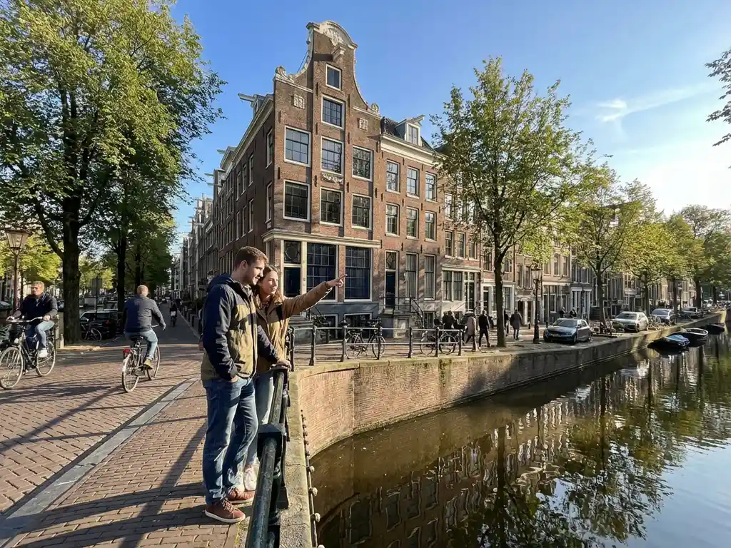 Jong stel op Amsterdamse gracht bekijkt historisch grachtenpand met trapgevel, vrouw wijst naar woning