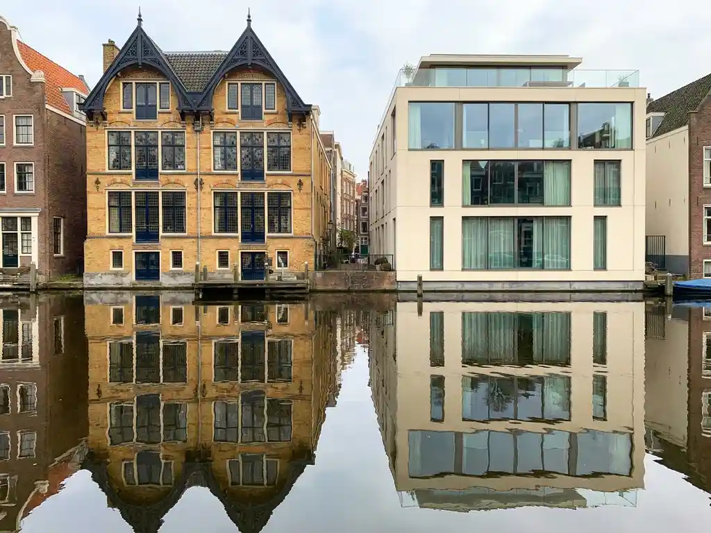 Historisch Amsterdams grachtenpand met 17e-eeuwse gevel naast modern glazen penthouse, weerspiegeld in kalm kanaalwater